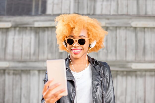 Ritratto di stile di vita di una donna africana in giacca di pelle che fa foto selfie con il telefono in piedi sullo sfondo della parete di legno