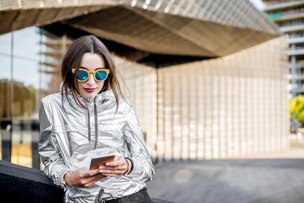 Ritratto di stile di vita di una donna elegante in giacca d'argento che utilizza il telefono all'aperto sullo sfondo architettonico moderno