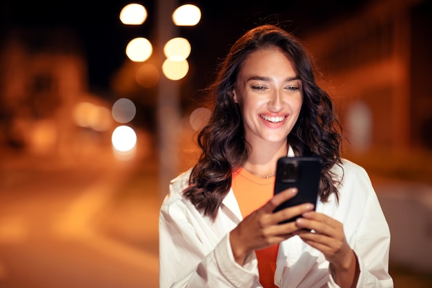 Ritratto di stile di vita di una donna felice che cammina nella città serale e usa il cellulare per inviare messaggi o navigare