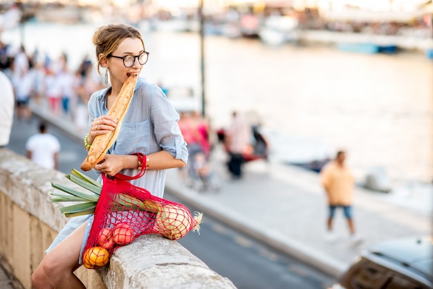 Ritratto di stile di vita di una giovane donna con borsa a rete piena di cibo fresco che mangia baguette all'aperto nella città vecchia