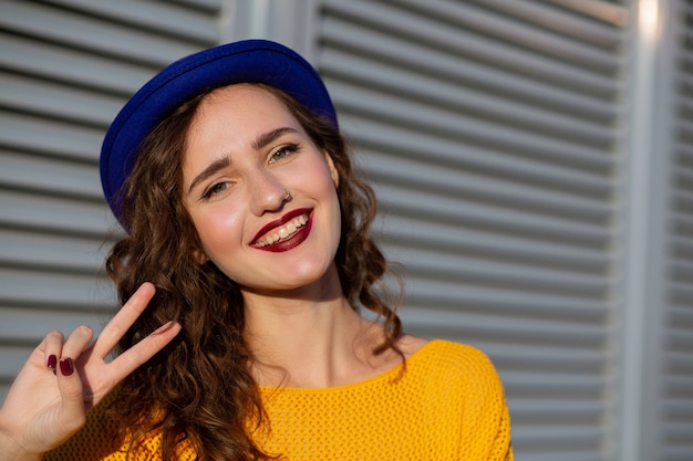 Ritratto di stile di vita: donna bruna eccitata in maglione giallo e cappello blu che mostra il segno della mano di vittoria. Spazio per il testo