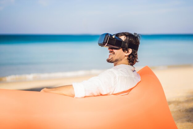 Ritratto di stile di vita estivo di uomo seduto sul divano gonfiabile arancione e utilizza cuffie per realtà virtuale sulla spiaggia dell'isola tropicale Rilassarsi e godersi la vita sul letto ad aria