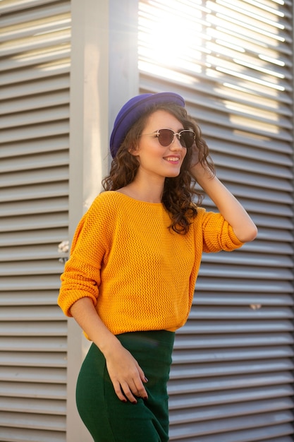 Ritratto di strada di un modello sorridente che indossa un cappello blu e un maglione lavorato a maglia giallo in posa sotto i raggi del sole