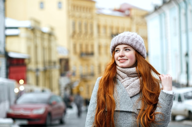 Ritratto di strada di una gioiosa ragazza rossa con i capelli lunghi che indossa abiti invernali caldi in posa per la strada. Spazio per il testo