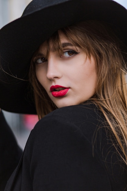 Ritratto di strada di una giovane modella lussuosa con labbra reg che indossa giacca e cappello neri
