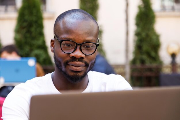 Ritratto di studente africano intelligente che indossa occhiali utilizzando il laptop mentre è seduto al bar