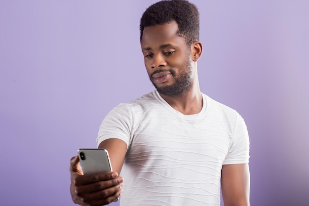 Ritratto di studente afroamericano amichevole attraente sicuro con capelli ricci corti e barba che indossa maglietta bianca che tiene il telefono cellulare prendendo selfie. Tecnologia e concetto di comunicazione.