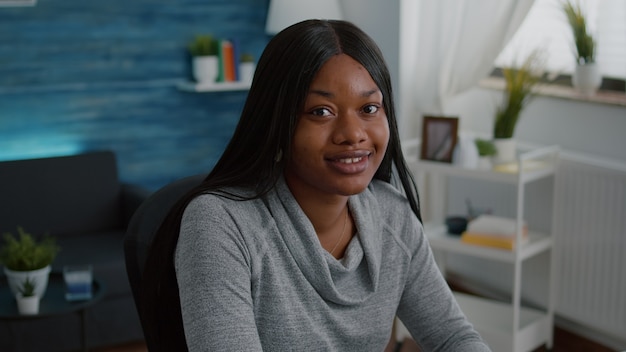 Ritratto di studente afroamericano seduto al tavolo della scrivania in soggiorno guardando la telecamera sorridendo durante l'istruzione a casa