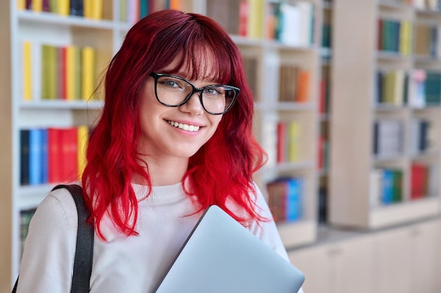 Ritratto di studentessa con zaino per laptop guardando la fotocamera in biblioteca Bella ragazza alla moda con i capelli tinti di rosso che indossa occhiali sorridente con i denti College University Education Youth