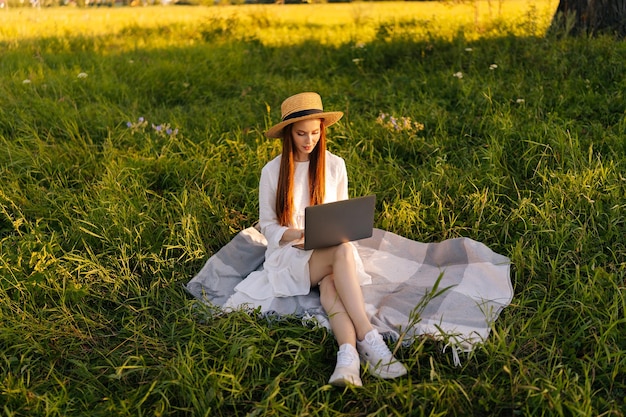 Ritratto di studentessa concentrata in cappello di paglia e abito bianco che studia sul computer portatile seduto sul bel prato di erba verde