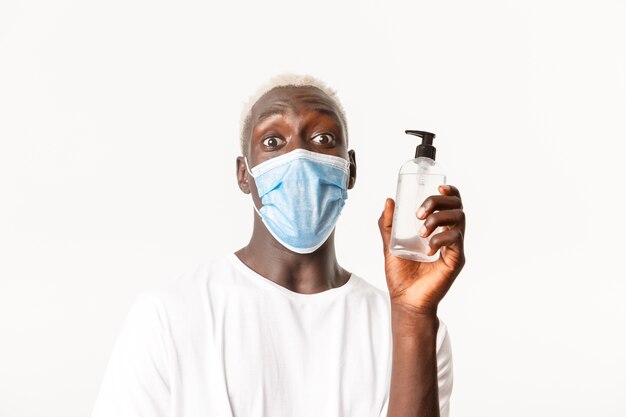 Ritratto di stupito ragazzo biondo afro-americano in mascherina medica, mostrando disinfettante per le mani e guardando eccitato