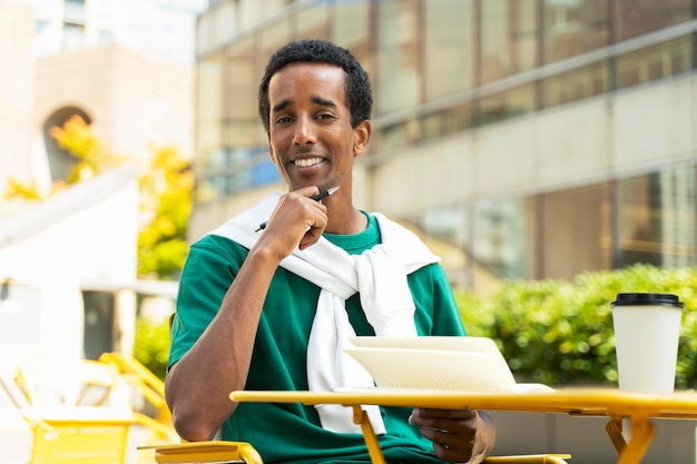 Ritratto di successo giovane attraente uomo d'affari afroamericano seduto sul posto di lavoro in un bar tenendo la penna prendendo appunti pausa caffè all'aperto sulla strada Uomo sorridente che indossa abiti casual