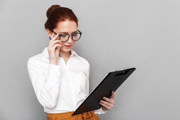 Ritratto di successo redhead imprenditrice 20s indossando occhiali tenendo appunti e sorridente in ufficio isolato su gray