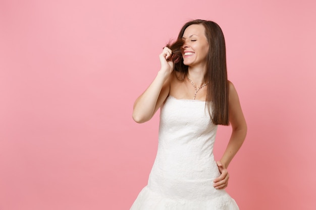 Ritratto di tenera donna sorridente con gli occhi chiusi in abito bianco in piedi tenendosi i capelli