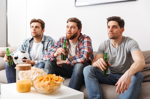 Ritratto di tre giovani uomini che guardano il calcio seduti a casa, bevendo birra e mangiando snack