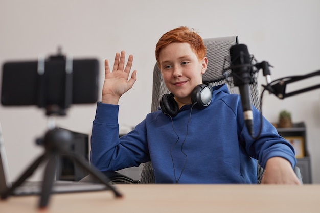 Ritratto di un adolescente dai capelli rossi che saluta la telecamera durante la registrazione di video o lo streaming online a casa, copia spazio