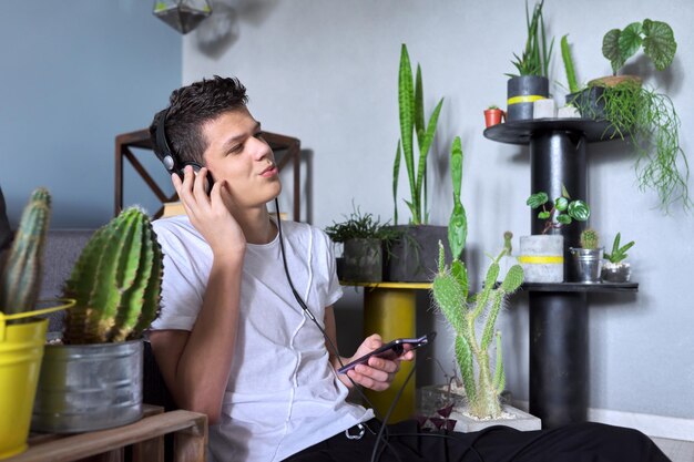 Ritratto di un adolescente di 16 anni in cuffia che ascolta musica, audiolibri, lezioni. Fondo in vaso dei cactus delle piante d'appartamento dell'interno domestico