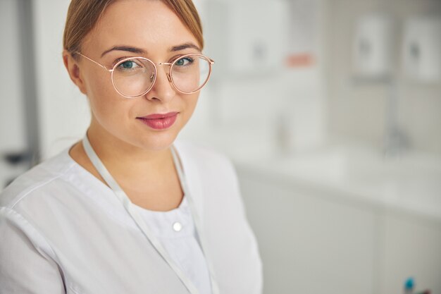 Ritratto di un'adorabile giovane donna bionda cosmetologa vestita con un camice da laboratorio che guarda avanti