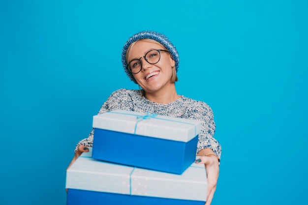 Ritratto di un'affascinante giovane donna dai capelli corti che dà giovani scatole regalo blu ridendo vestita in abiti blu contro il muro blu dello studio.