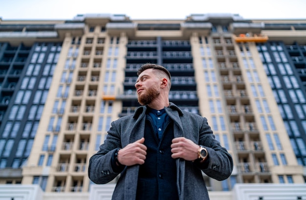 Ritratto di un affascinante giovane uomo d'affari vestito con giacca e cappotto. In piedi fuori da un edificio moderno.