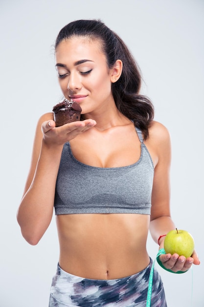Ritratto di un'affascinante ragazza fitness scegliendo tra mela e torta isolata su un muro bianco