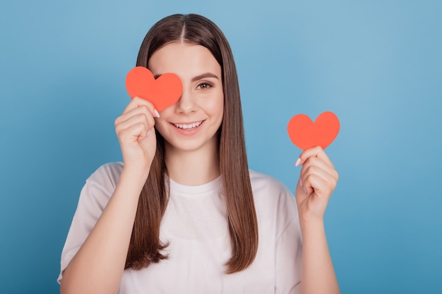 Ritratto di un'affascinante ragazza timida e giocosa che tiene due piccoli occhi rossi a forma di cuore su sfondo blu