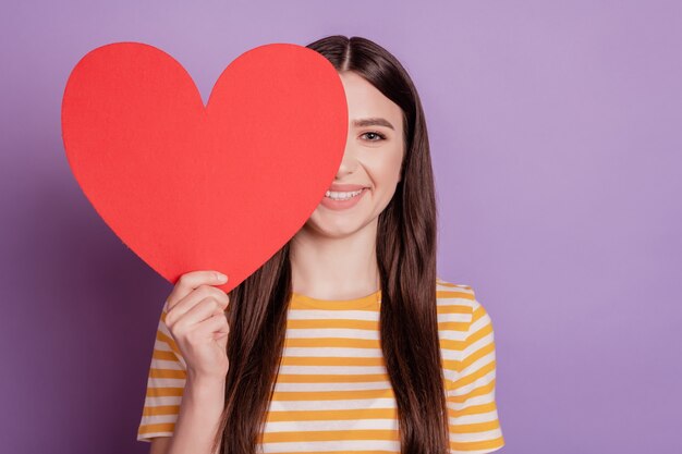 Ritratto di un'affascinante signora splendida che tiene l'occhio di copertina del cuore di carta rossa su sfondo viola