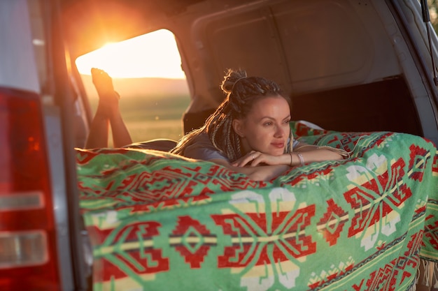 Ritratto di un'allegra ragazza hippie che giace su una coperta luminosa all'interno di un minivan e guarda la natura...