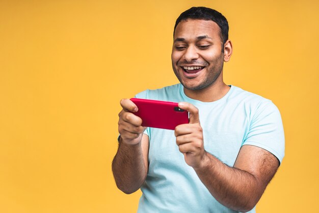 Ritratto di un allegro giovane africano nero indiano uomo vestito con giochi casuali sul telefono cellulare isolato su sfondo giallo.