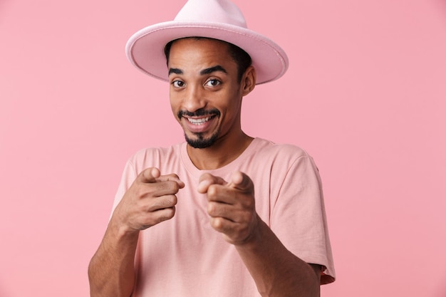 Ritratto di un allegro uomo afroamericano che indossa un cappello sorridente e punta le dita isolato su rosa