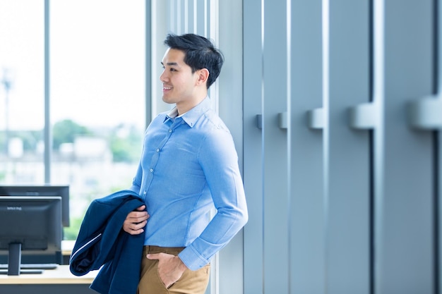 Ritratto di un allegro vincitore sentimento di successo bel giovane uomo d'affari asiatico indossare un tailleur di uomo in giacca blu e camicia blu alla finestra Sullo sfondo della stanza dell'ufficio