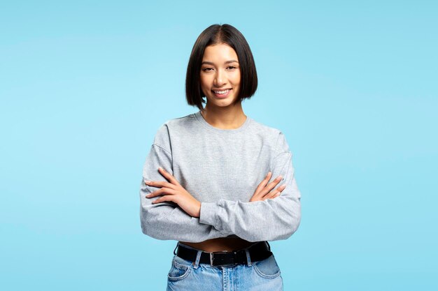 Ritratto di un'attraente donna asiatica che indossa abiti casuali eleganti con le braccia incrociate che guarda la telecamera in piedi isolata su uno sfondo blu