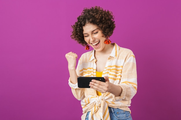 Ritratto di un'attraente giovane donna allegra con i capelli ricci bruna in piedi isolata sul muro viola, giocando al telefono cellulare, celebrando il successo