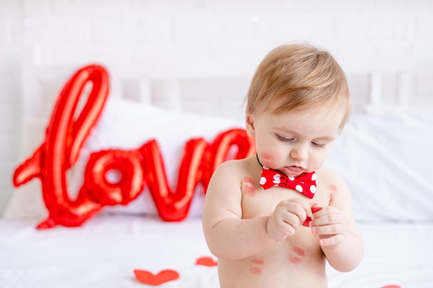 Ritratto di un bambino biondo seduto su un letto in pannolini con baci dal rossetto rosso a casa e sorridente tra i cuori e un grande palloncino con la scritta ama il concetto di San Valentino