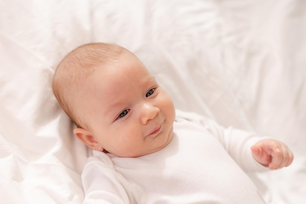 Ritratto di un bambino carino con gli occhi azzurri in un body bianco sdraiato sulla schiena su lenzuola bianche