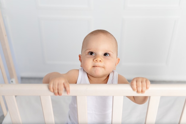 Ritratto di un bambino sorridente 8 mesi in piedi in una culla nella stanza dei bambini in abiti bianchi