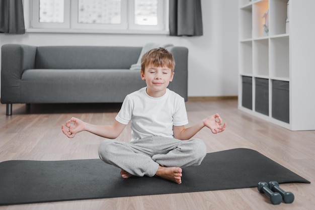 Ritratto di un bambino sorridente durante una sessione di meditazione yoga, seduto nell'esercizio Padmasana, posa del loto con Namaste, che si allena indossando abiti da casa, gli occhi chiusi.