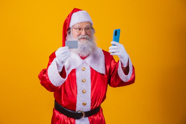 Ritratto di un bel Babbo Natale sbalordito e barbuto che tiene in mano un dispositivo di carta di plastica negozio di app web acquistare ordine consegna pagare pagamento wireless isolato su sfondo giallo