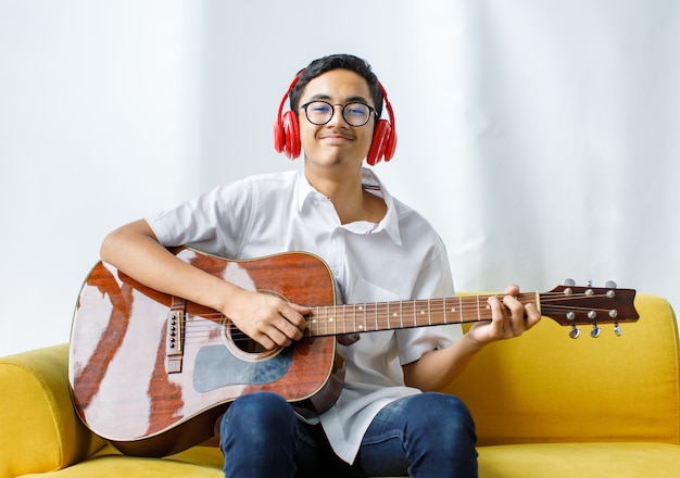 Ritratto di un bel giovane adolescente maschio che si diverte a suonare la chitarra acustica e indossa una cuffia rossa. Chitarrista junior in abiti bianchi seduto sul divano e in possesso di uno strumento