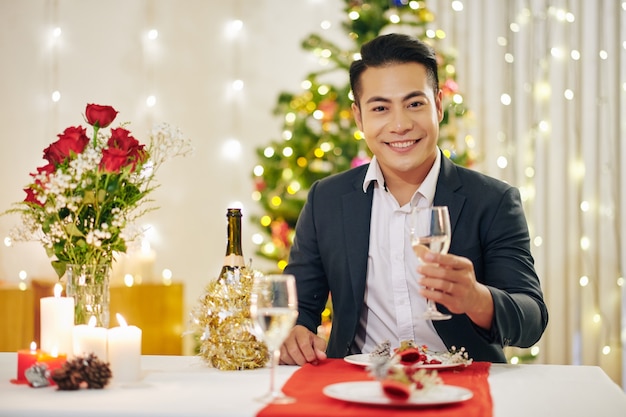 Ritratto di un bel giovane felice seduto al tavolo della cena di Natale con un bicchiere di champagne