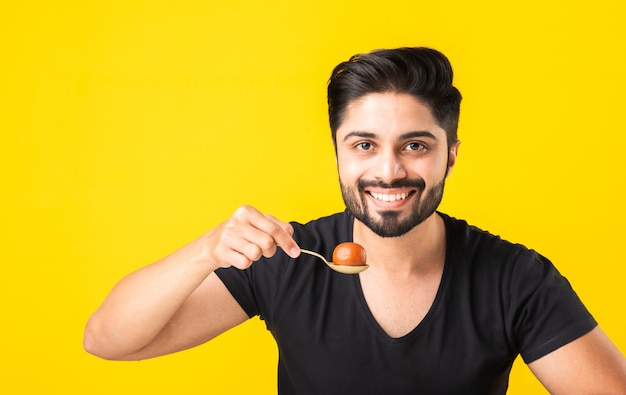 Ritratto di un bel giovane indiano che mangia dolce Gulab Jamun su sfondo giallo