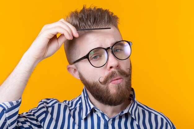 Ritratto di un bel giovane maschio hipster con baffi e barba che pettina i capelli contro un muro giallo. Concetto di barbiere e cura di sé. Posto per promo