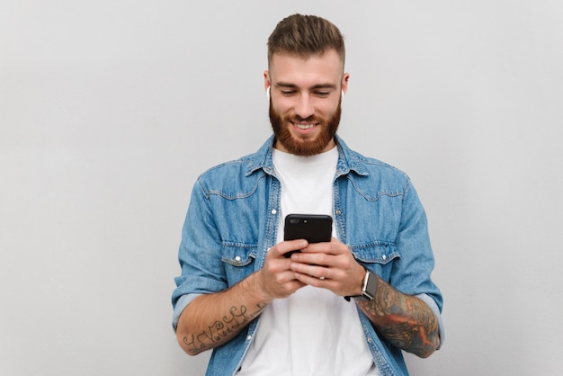 Ritratto di un bel giovane sorridente che indossa abiti casual in piedi isolato su un muro grigio, ascoltando musica con auricolari wireless, tenendo il telefono cellulare