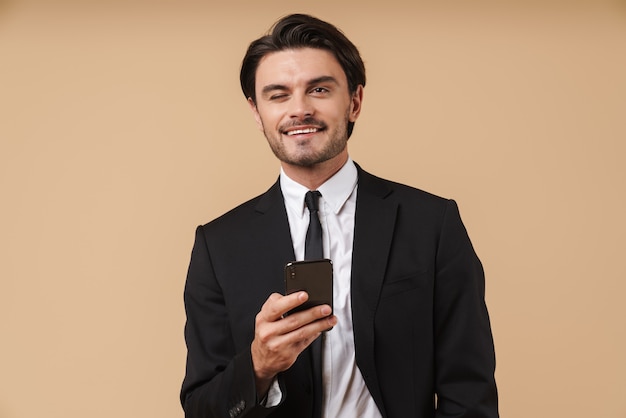 Ritratto di un bel giovane uomo d'affari sorridente che indossa un abito in piedi isolato su un muro beige, usando il telefono cellulare