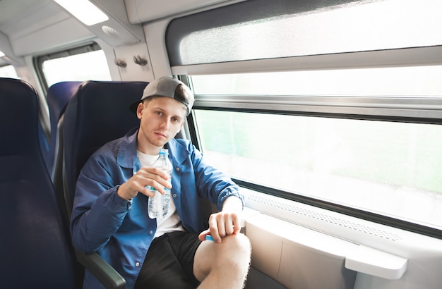 Ritratto di un bel giovane uomo seduto in un treno vicino a una finestra con una bottiglia di acqua nelle sue mani