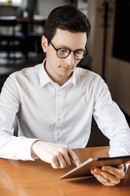 Ritratto di un bel giovane uomo utilizzando una tavoletta mentre è seduto a un comò scrivania in camicia bianca con gli occhiali.