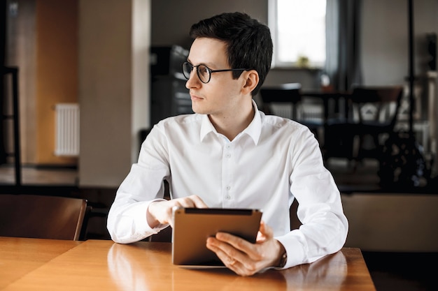 Ritratto di un bel manager adulto che indossa una camicia bianca e occhiali che guarda lontano mentre si tiene un tablet seduto alla scrivania in un coffee shop.