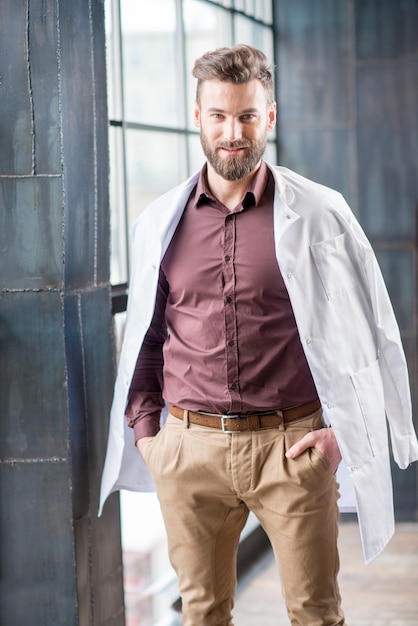Ritratto di un bel medico fiducioso con abito medico in piedi vicino alla finestra nella moderna clinica o ufficio interno scuro