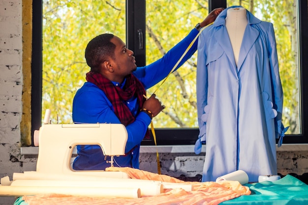 Ritratto di un bell'uomo africano sarta con macchina da cucireAfrio uomo americano elegante designer che lavora in sartoria manichino coattable nastro di misurazione in camera contro la finestra sera