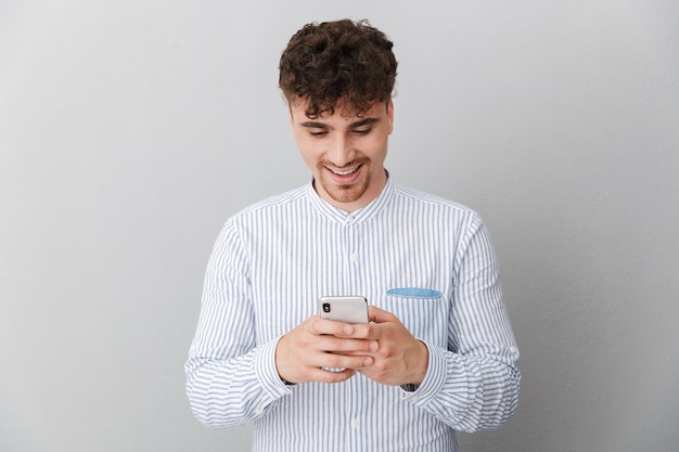 Ritratto di un bell'uomo allegro vestito con una camicia che sorride mentre tiene e usa lo smartphone isolato su un muro grigio